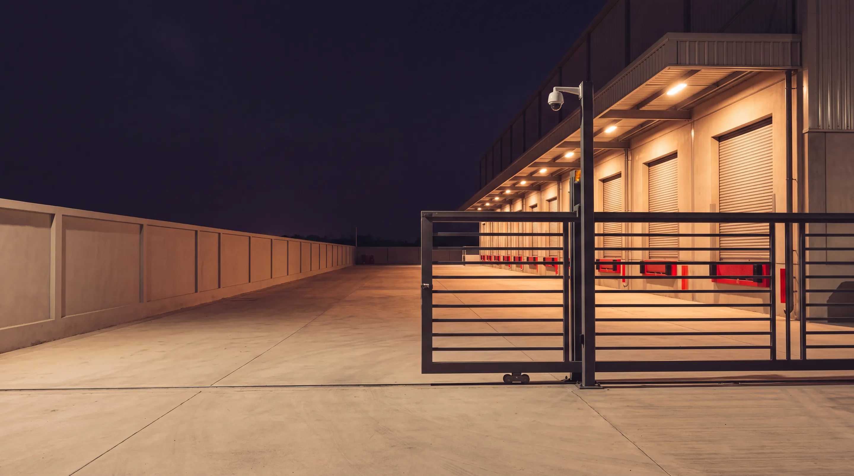 A warehouse loading area behind a sliding gate and boundary wall at night, with a camera on a pole